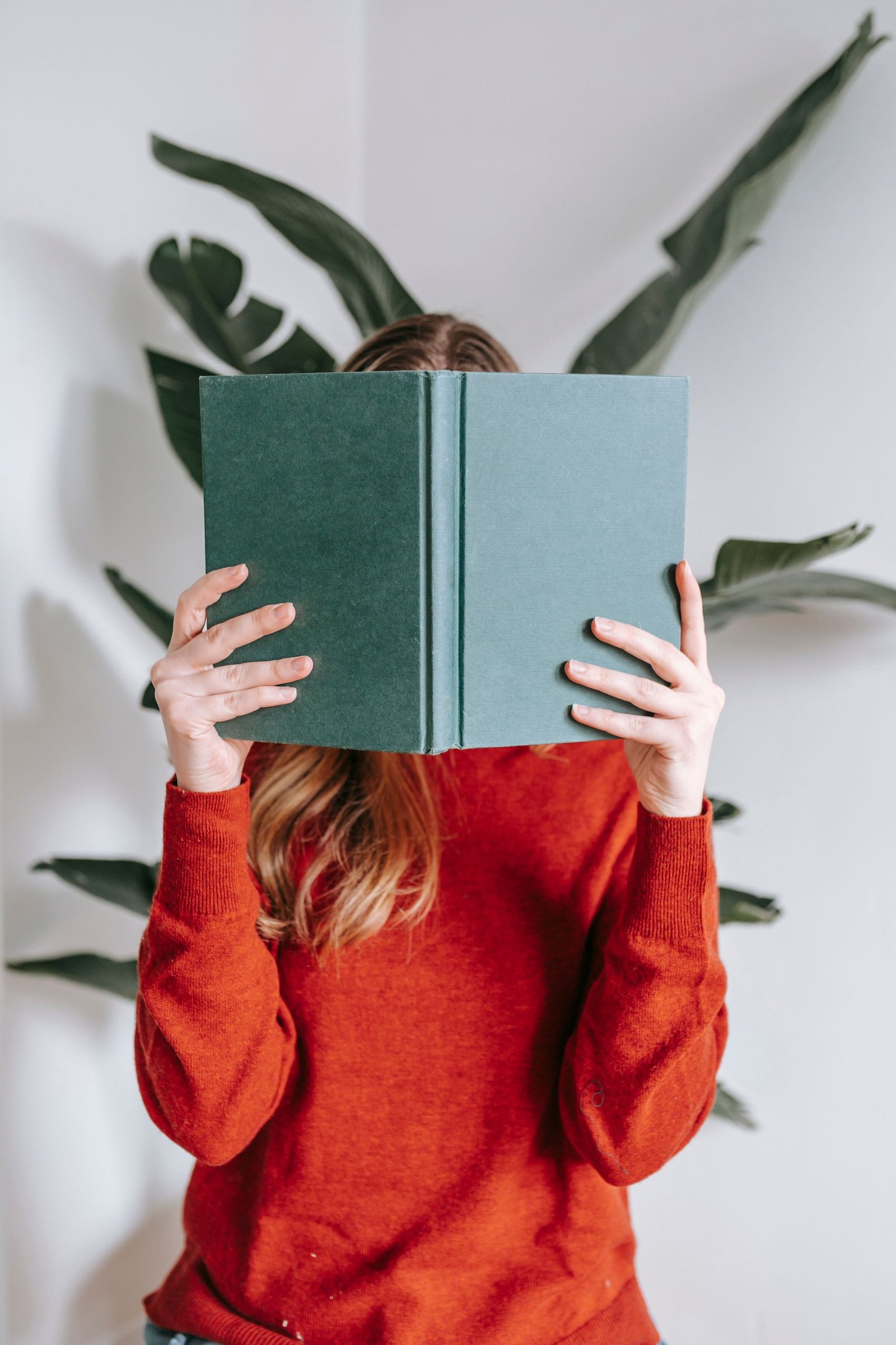 A woman indoors hiding face behind a green book, wearing a red sweater against a white wall with green leaves.